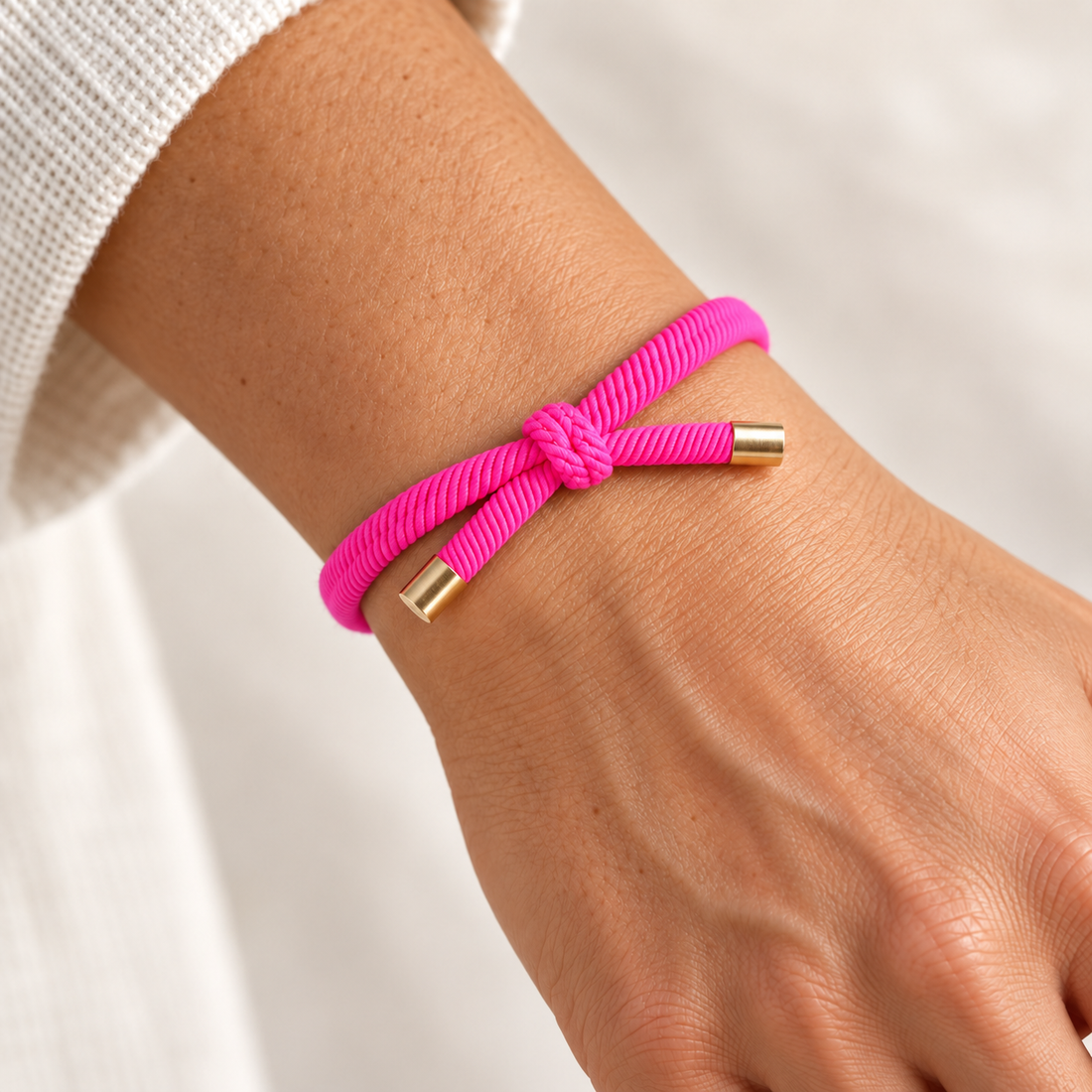 FUCHSIA Hårelastik og armbånd båret på et håndled med synlig knude og guldfarvede endestykker.