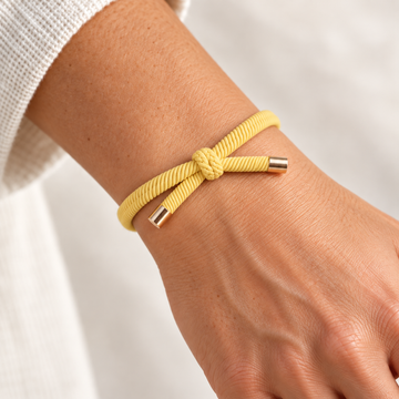GUL Hårelastik og armbånd båret på et håndled med synlig knude og guldfarvede endestykker.