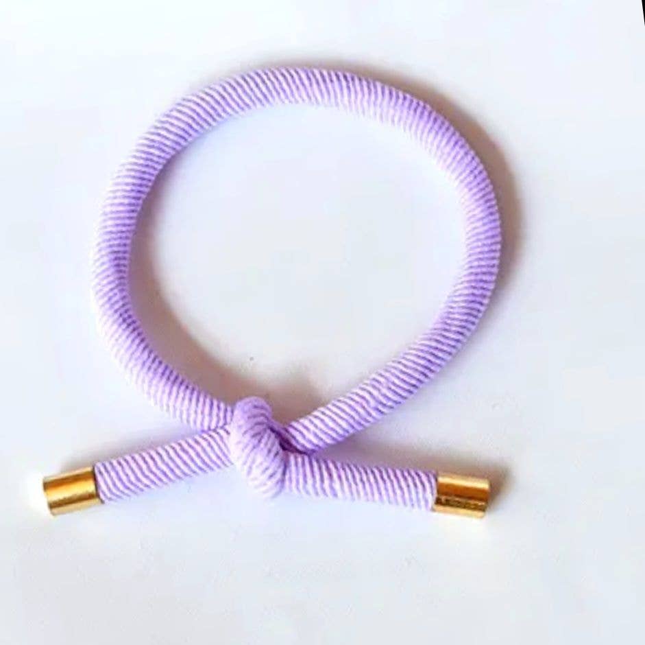 LILLA Hårelastik og armbånd med dekorativ knude og guldfarvede endestykker på lys baggrund.
