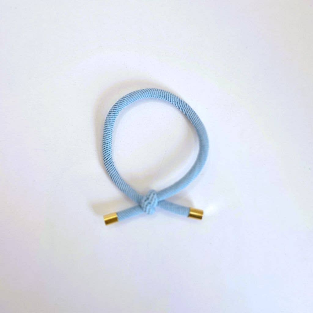 LYSEBLÅ Hårelastik og armbånd med knude og guldfarvede endestykker på lys baggrund.